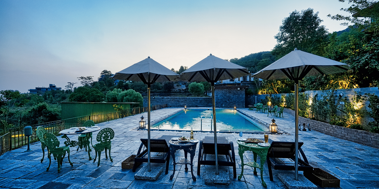 An image of outdoor pool with lounge chairs and umbrellas, surrounded by greenery - Atman, Dharamshala.