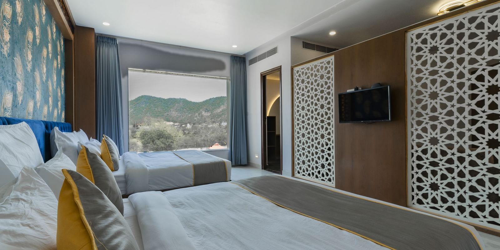 An interior shot of a room with a bed and a decorative partition and a mountain view is visible through a window in the background - Bamboo Saa Mulberry Resort, Pushkar