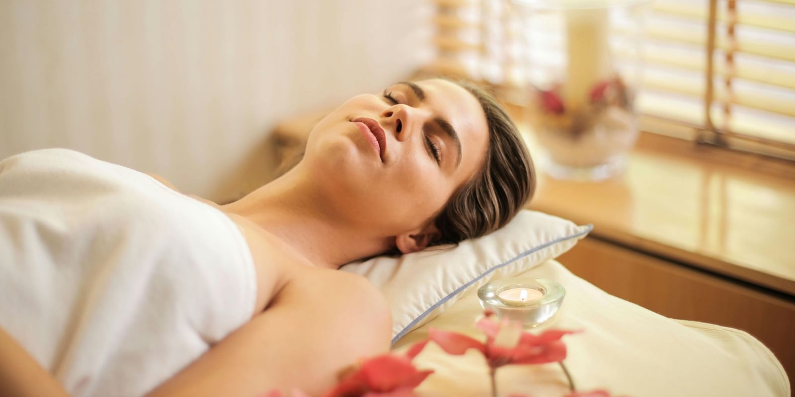A person lies relaxed on a spa bed, eyes closed, with a towel draped over them and flower petals nearby