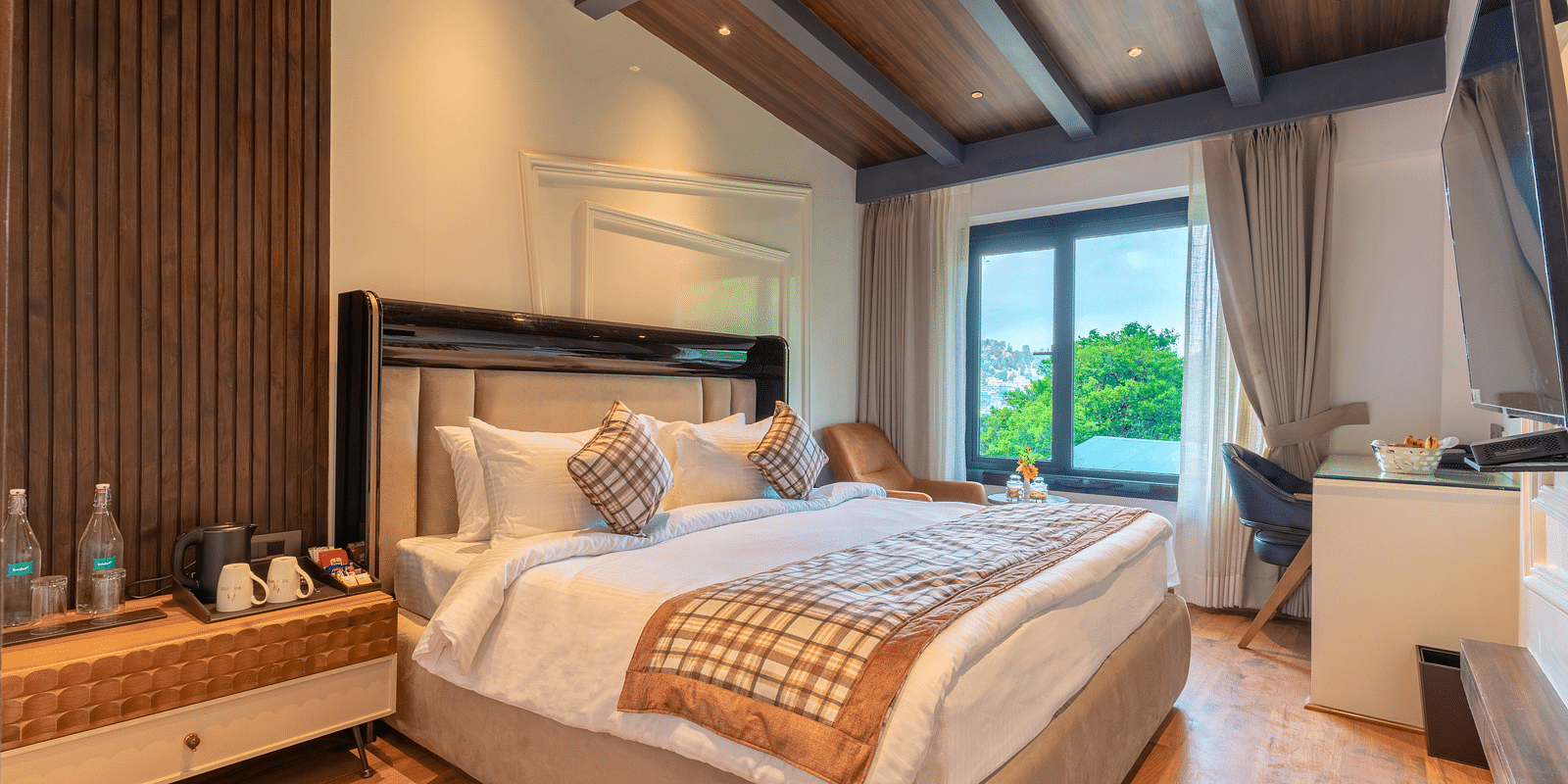 An image of a bedroom with king size bed, bedside table where bottles are kept, a chair and table, and a flat TV screen - Bedzzz Xclusiv Ikshana Luxe, Nainital