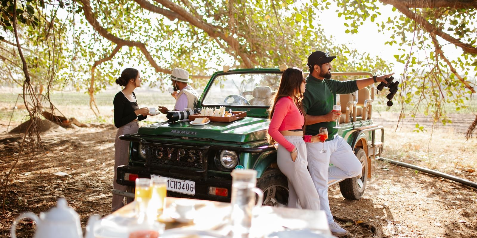 Four people standing beside a jeep which is standing before a breakfast table, surrounded by trees -Daksh Eden Greenz, Sasan Gir