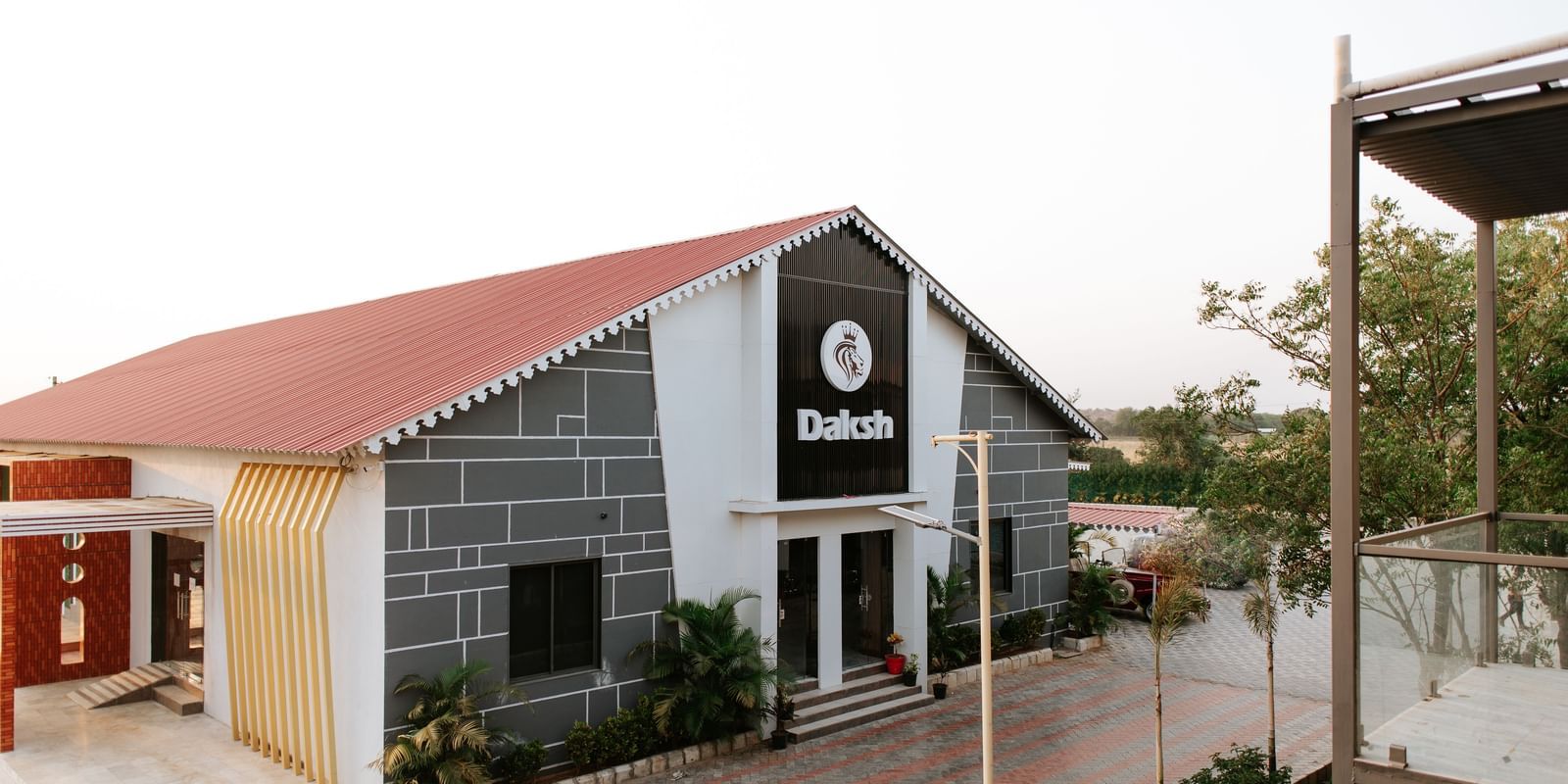 A side angle shot of a the exterior of Daksh Eden Greenz, Sasan Gir on a bright day, surrounded by trees and spacious walkways.