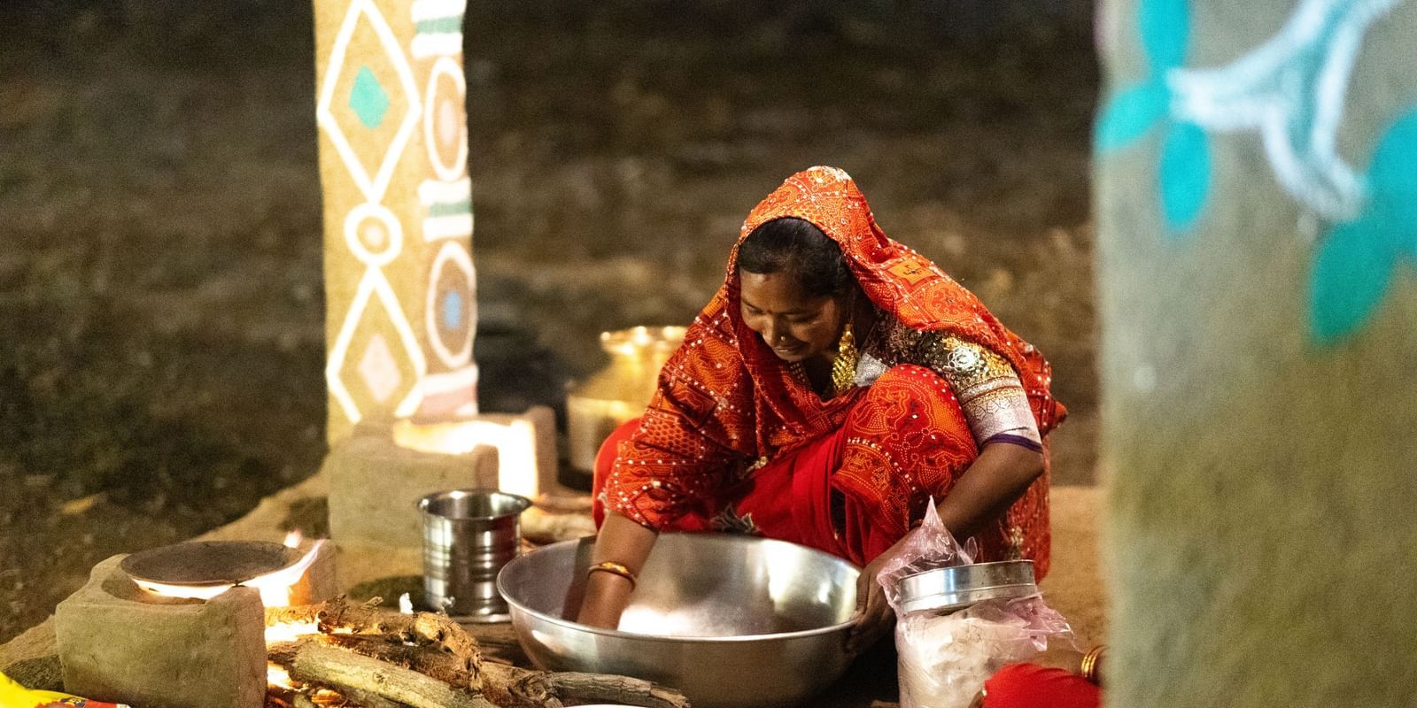 A woman preparing food in large steel utensils while seated on the ground in a traditionally built kitchen at Daksh Edden Greenz, Sasan Gir.
