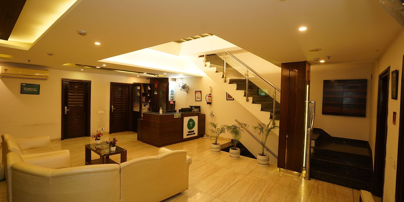 A hotel lobby with a curved white sofa, a small coffee table, a staircase leading to an upper level, and a reception desk in the background - Diamond Hotels