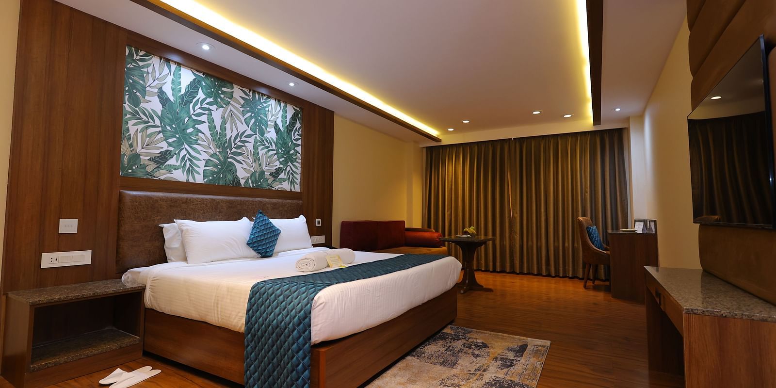 Side angle image of a Suite at Amar Hotel with a bed, table, TV and a painting