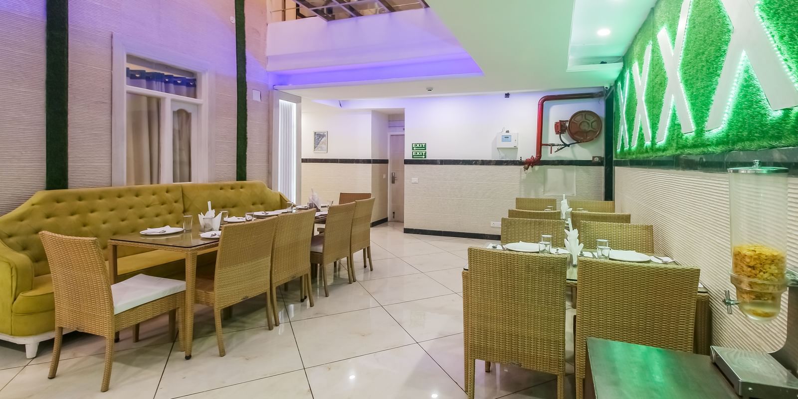 A restaurant interior with a long tufted sofa, wooden tables and chairs, a decorative wall design, and a tiled floor, lit by soft lighting. - Hotel Diamond Plaza, Chandigarh.
