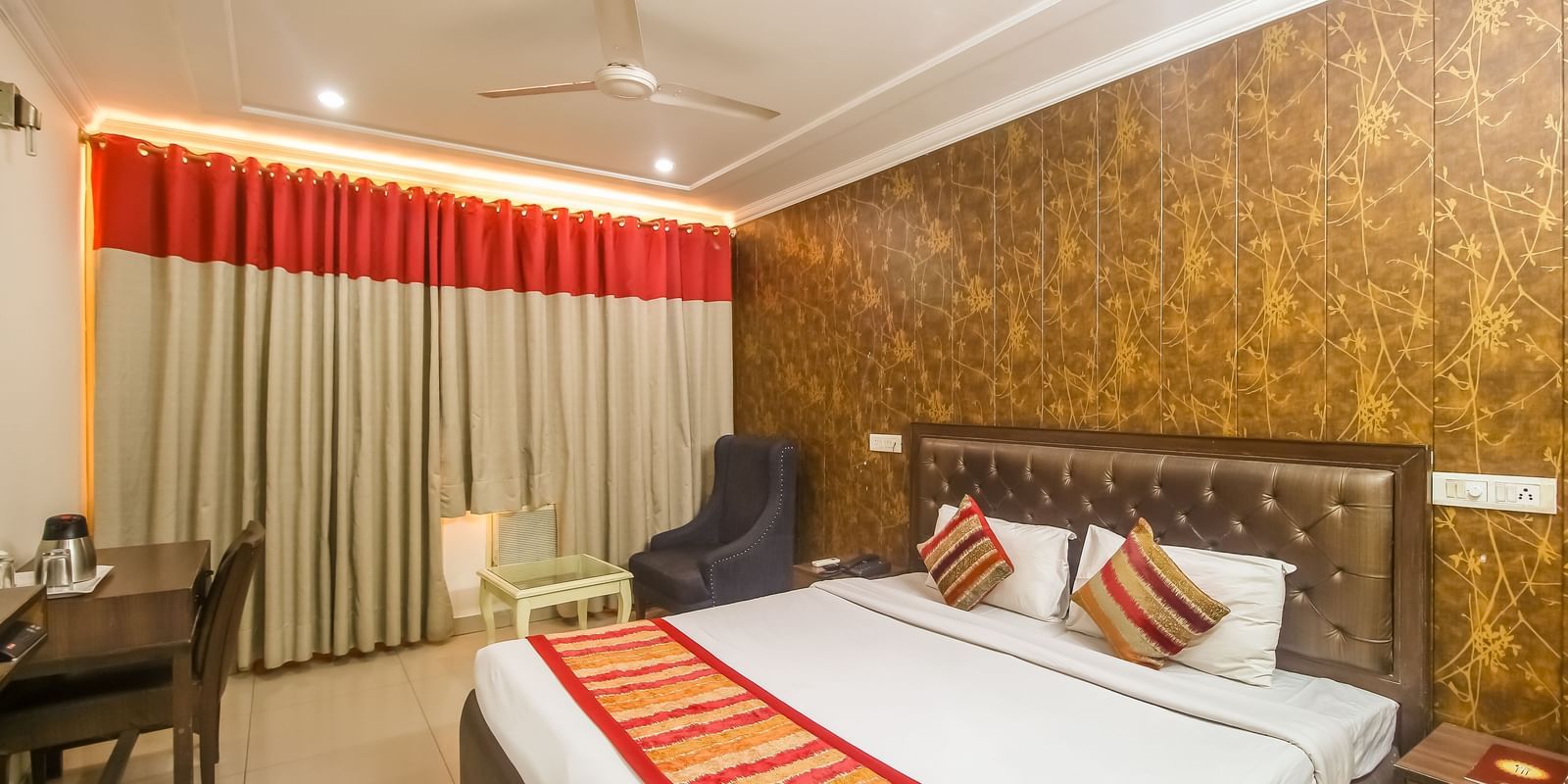A hotel room with a textured accent wall, a plush bed with decorative pillows and a runner, a dark armchair, and a desk, illuminated by warm lighting. - Hotel Diamond Plaza, Chandigarh.