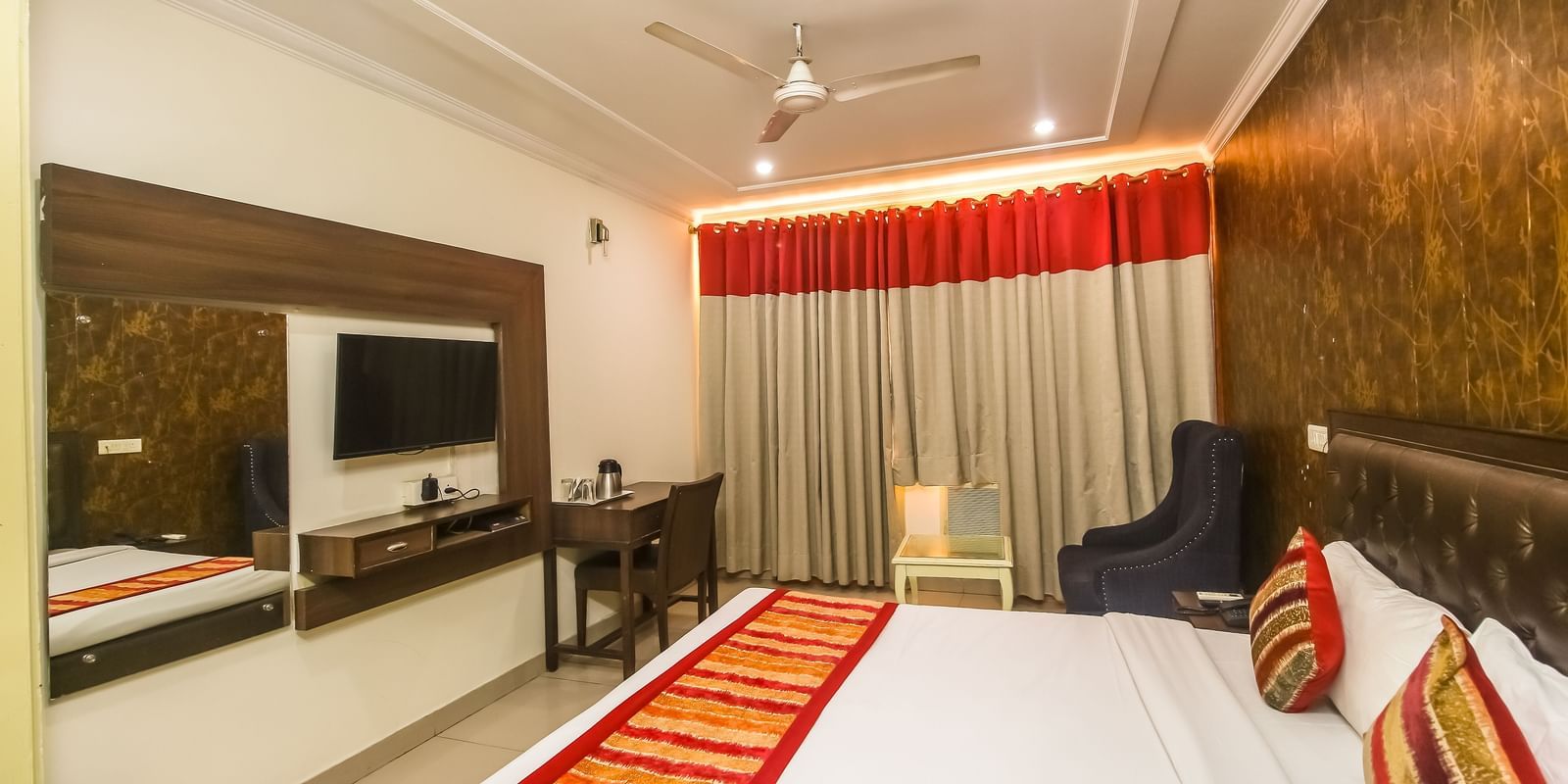A hotel room with a plush bed featuring a patterned runner, a TV, a desk, a dark armchair, and a textured accent wall, lit by soft lighting. - Hotel Diamond Plaza, Chandigarh.