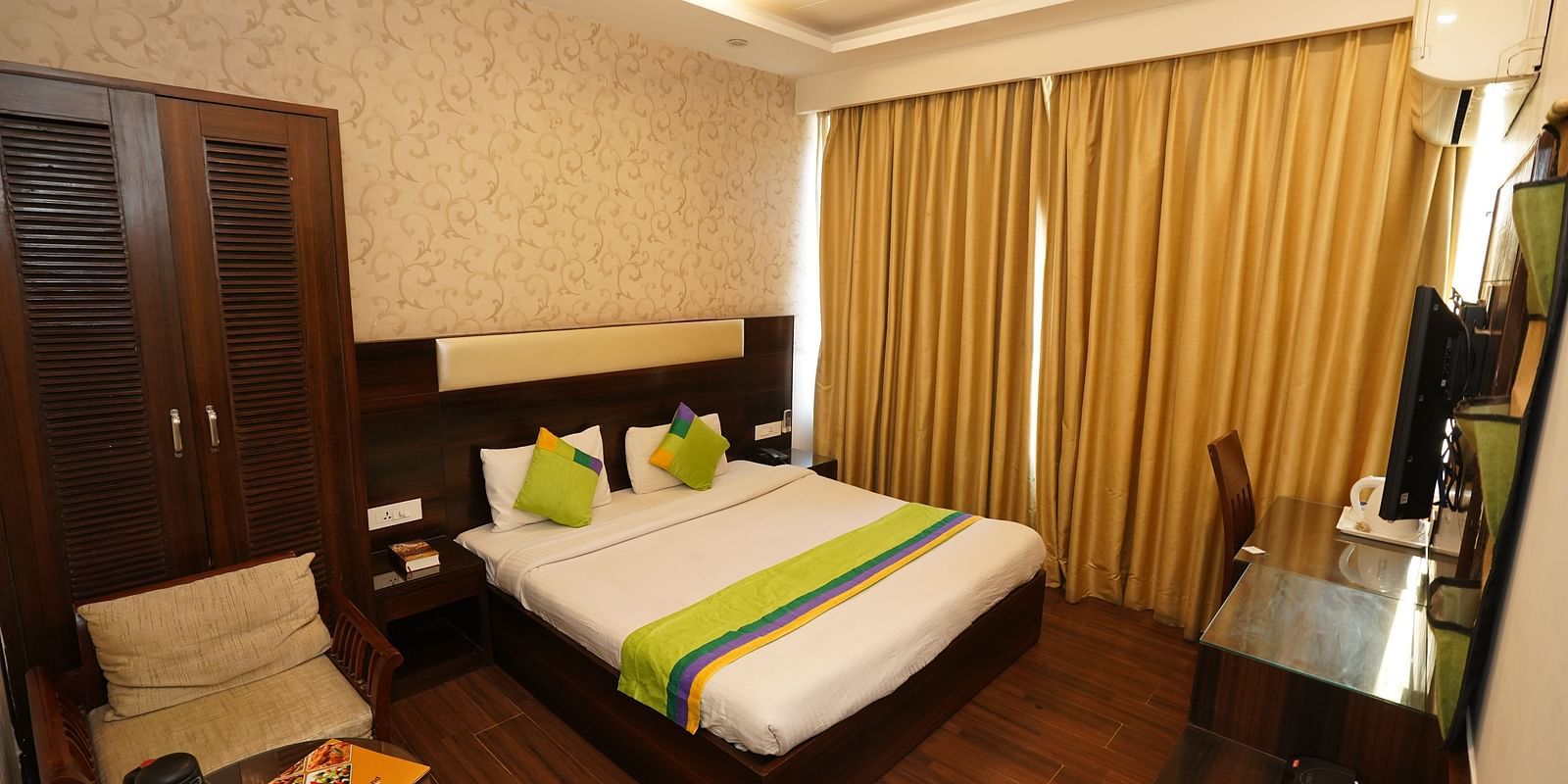 Deluxe Room featuring a king size bed with green accents, a wooden headboard, bedside tables, curtains, a desk, and a chair.- Hotel Dreamland, Chandigarh