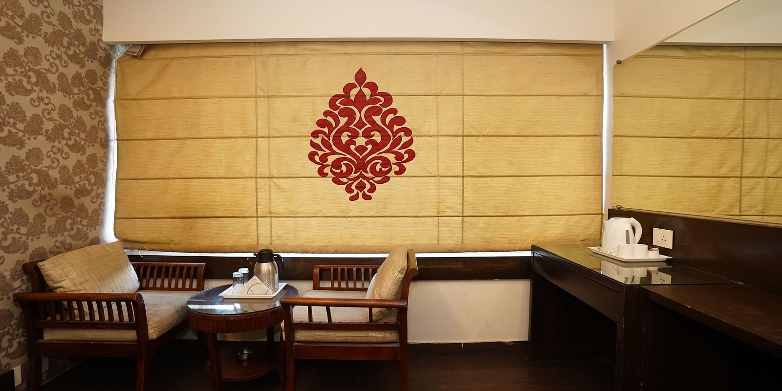 Deluxe Room featuring a seating area with chairs and a small table, a window covered by a decorative blind with a red pattern, and a desk with a kettle, set against a backdrop of textured walls and warm lighting.- Hotel Dreamland, Chandigarh