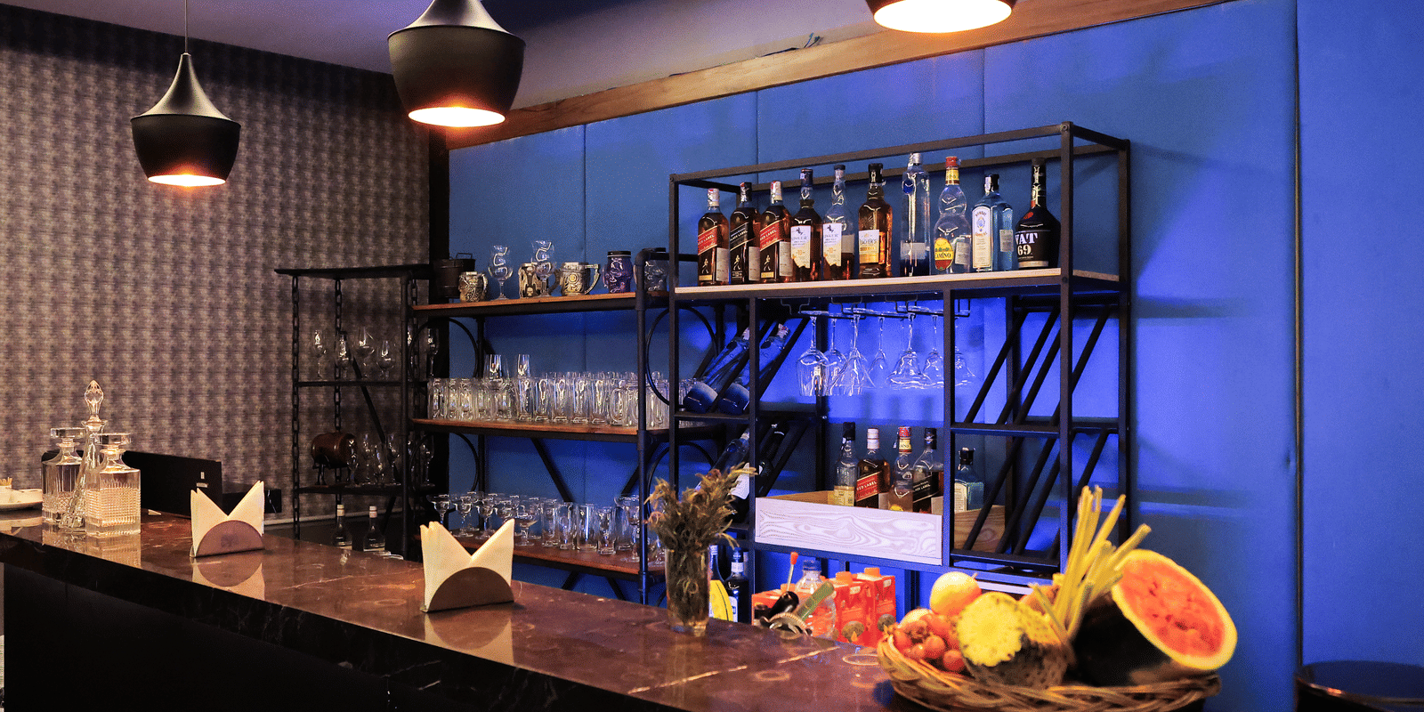 Stylish bar counter with fruit and glasses, a black metal shelving unit, and a blue accent wall with lighting at Hotel Hukam's Lalit Mahal.