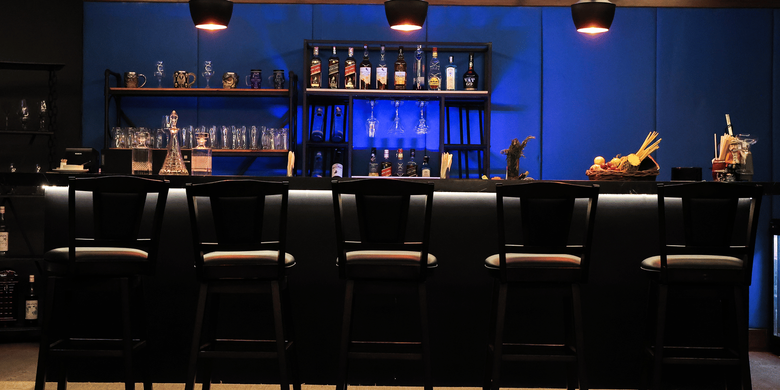Wide view of the dark, sleek bar with a blue accent wall, shelving full of bottles, and black leather bar stools at Hotel Hukam's Lalit Mahal.