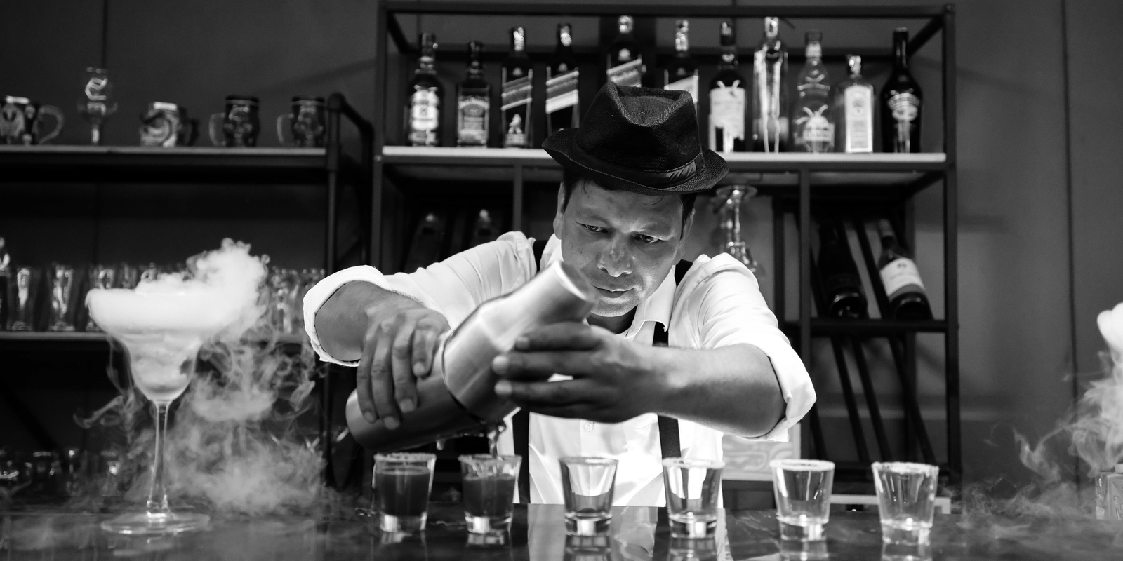 Monochrome image of a bartender in a hat creating a smoky effect over cocktails for a dramatic presentation at Hotel Hukam's Lalit Mahal.