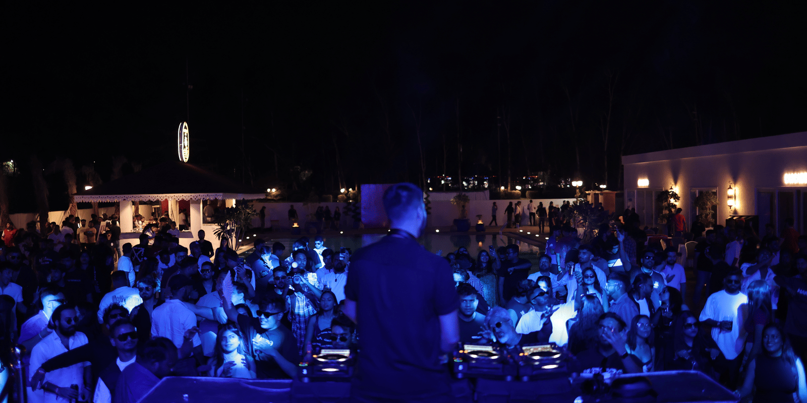 Crowded night-time event with people dancing, focused on a DJ console under bright blue stage lighting at Hotel Hukam's Lalit Mahal.