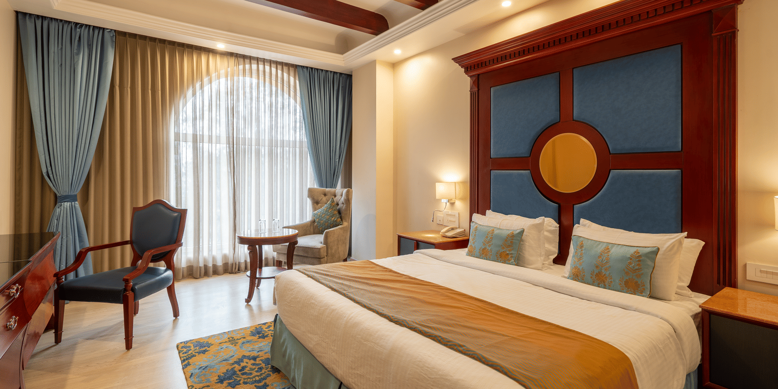 Luxurious hotel room with a large bed, blue and gold decor, a grand headboard, and arched window with sheer curtains at Hotel Hukam's Lalit Mahal.