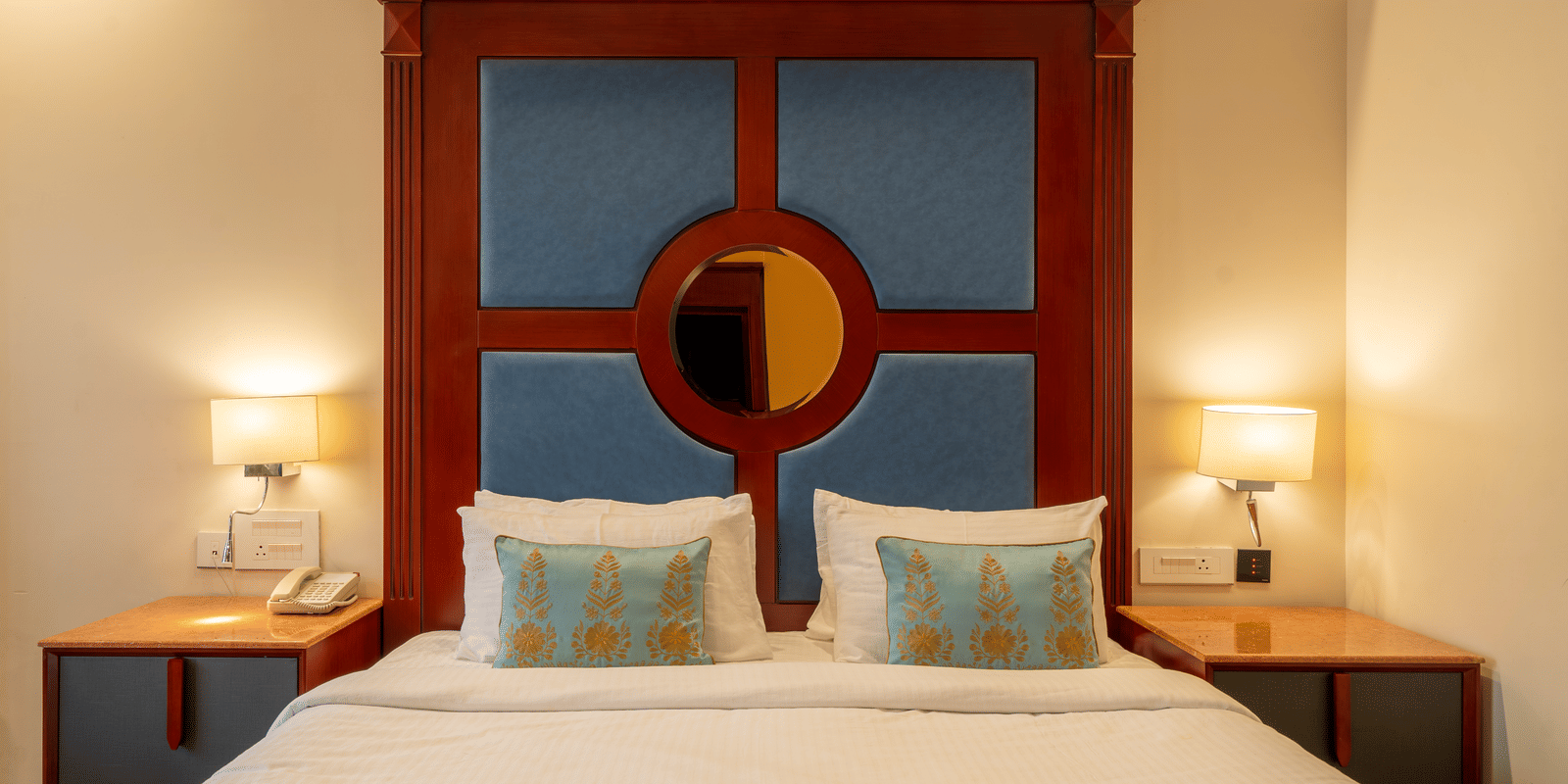 Close-up of a double bed with pillows, flanked by lamps and a striking blue and mahogany headboard design at Hotel Hukam's Lalit Mahal.