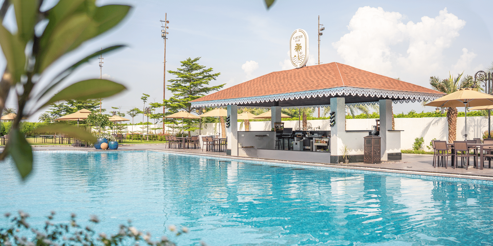 Sunny daytime view of the swimming pool with a red-roofed bar or pavilion and lush greenery in the foreground at Hotel Hukam's Lalit Mahal.