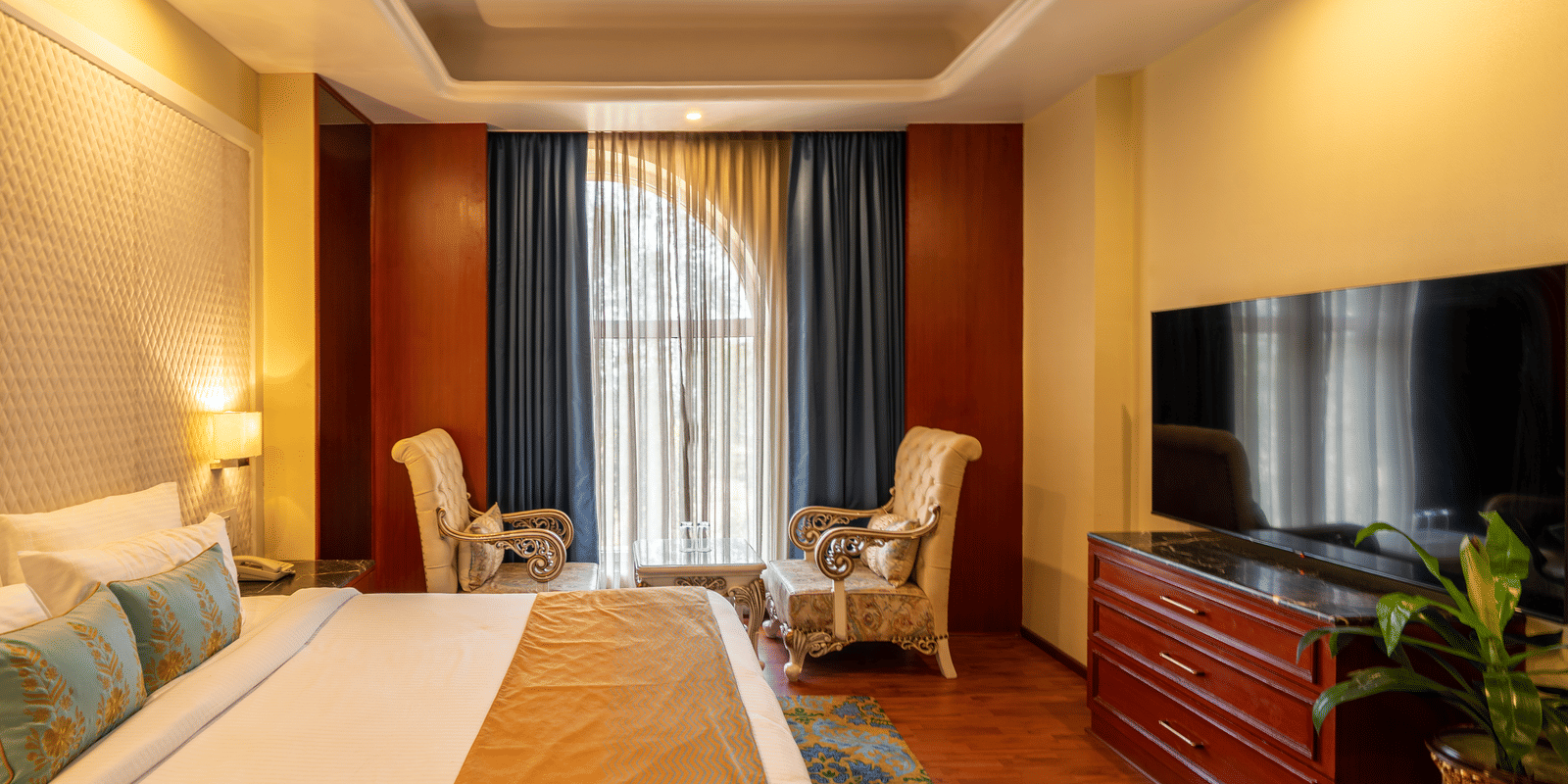 Hotel bedroom featuring a television above a wooden chest, comfortable seating, and a large bed at Hotel Hukam's Lalit Mahal.