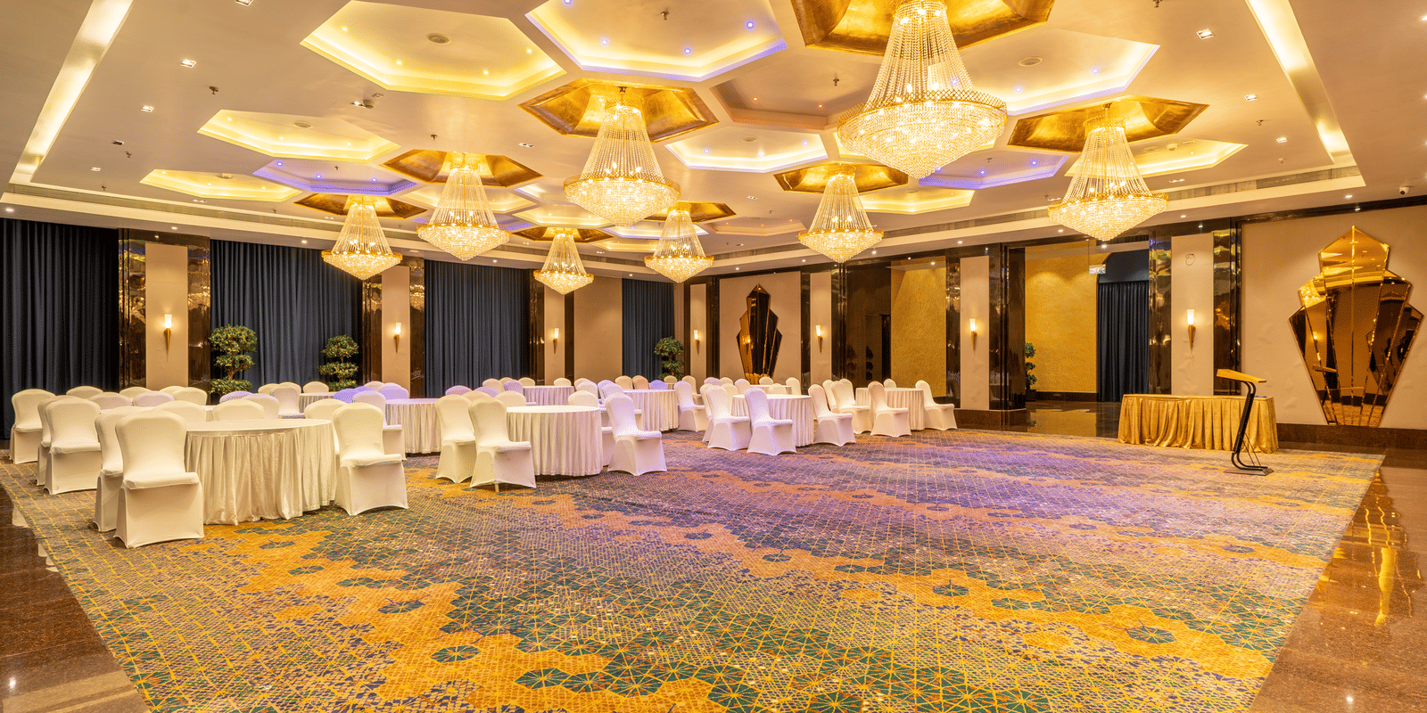 Wide view of a long banquet table covered in white cloth, set up under ornate gold and white hexagonal chandeliers at Hotel Hukam's Lalit Mahal.