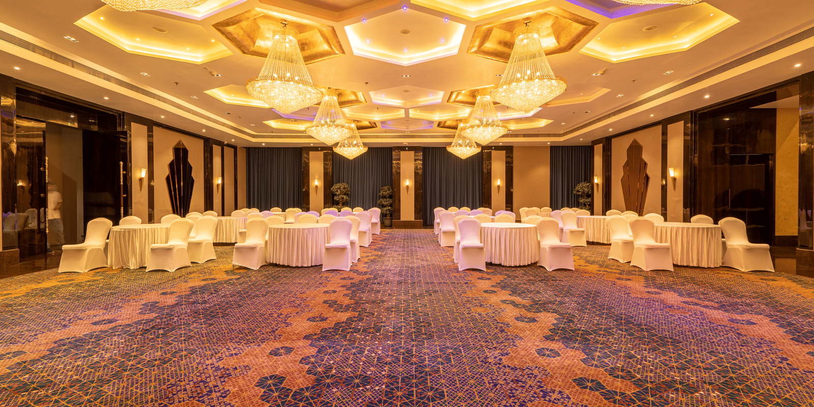 Grand banquet hall with a patterned carpet, dark walls, and stunning hexagonal crystal chandeliers on the ceiling at Hotel Hukam's Lalit Mahal.
