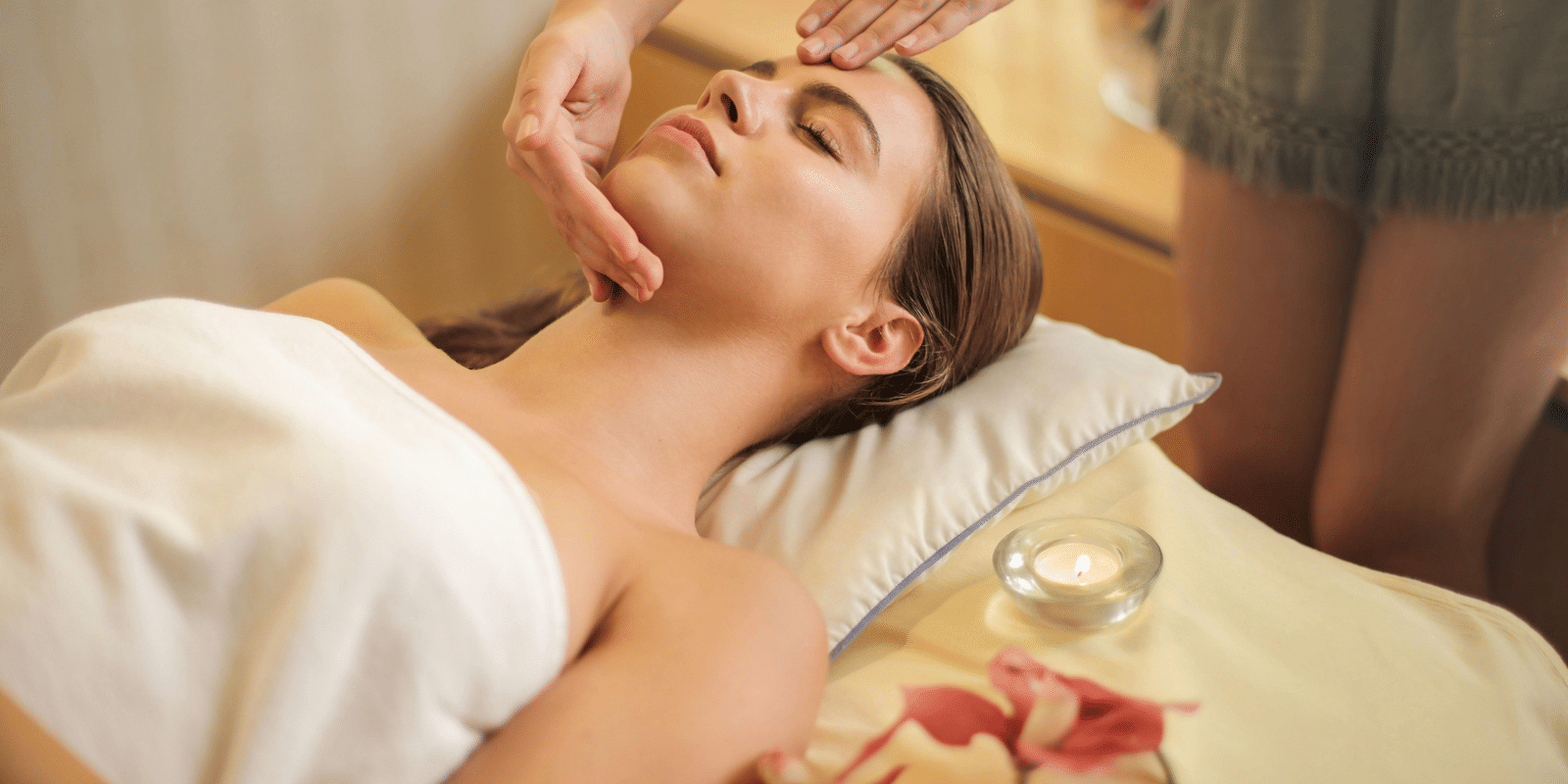 A woman relaxing during a facial massage at a spa, lying down with rose petals nearby and a white towel wrapped around her.