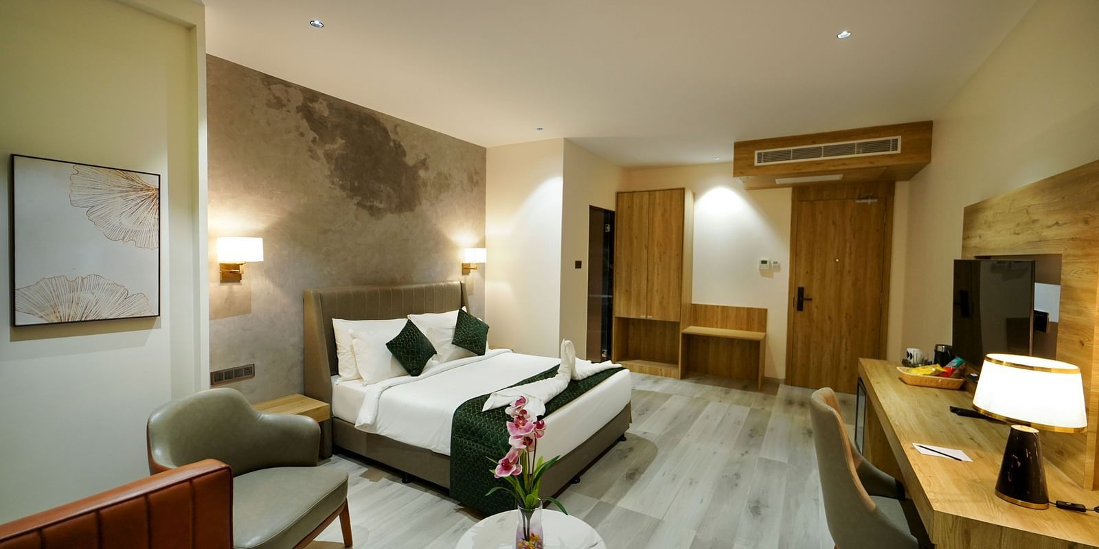 A spacious hotel room includes a bed with pillows, a seating area with chairs and a small table with flowers, and a desk area - Hotel Park Inn, Nellore.