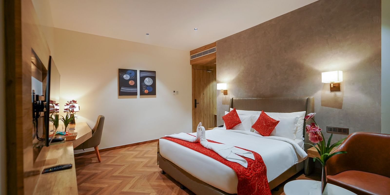 A hotel room includes a bed with pillows and folded linens, a desk area with a chair, a round table with flowers, and artwork on the wall - Hotel Park Inn, Nellore.