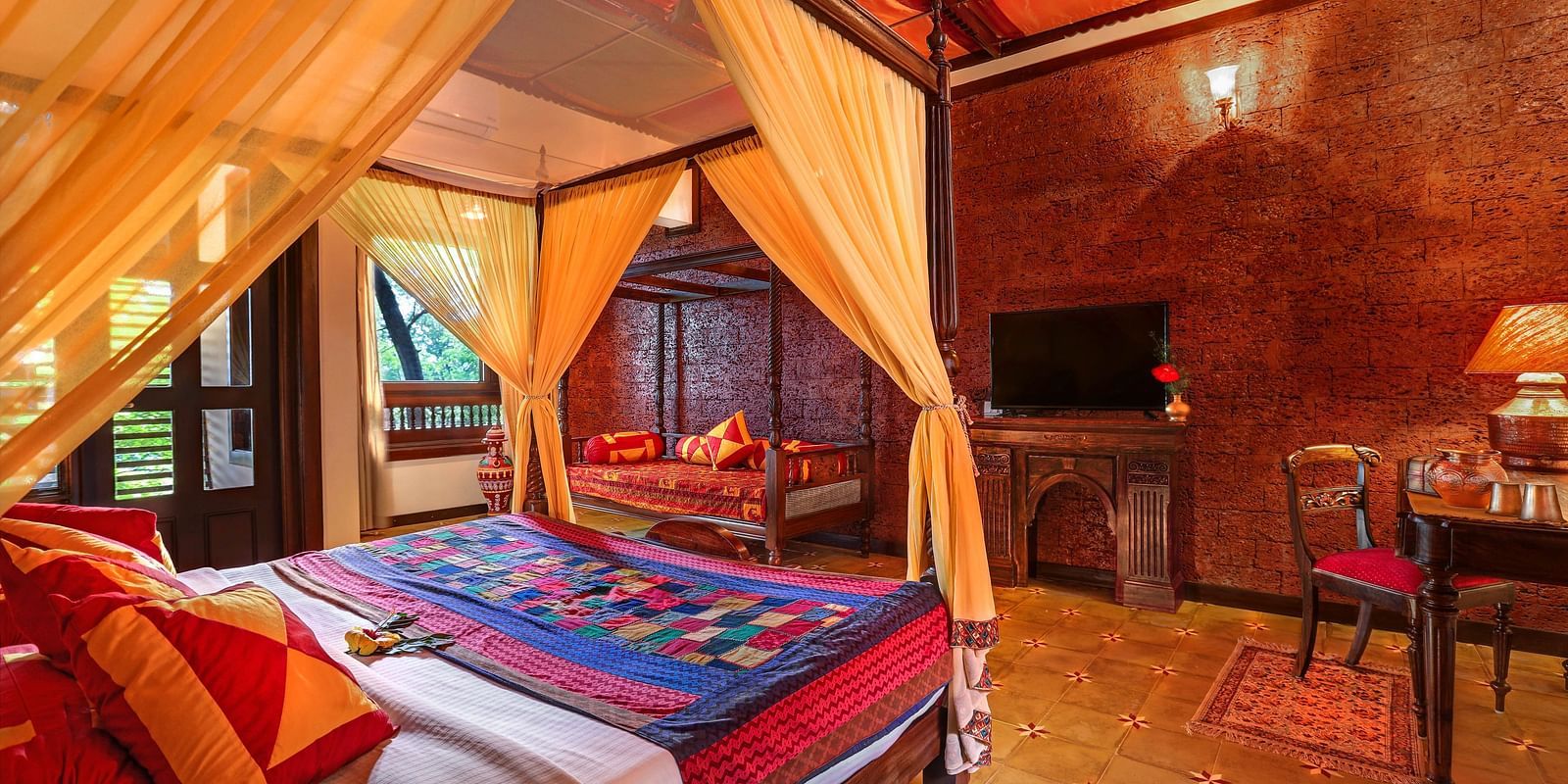 An image of a master suite bedroom with poster bed, windows, flat TV screen, wooden table, lamp, and many more - Hoysala Village Resort, Hassan