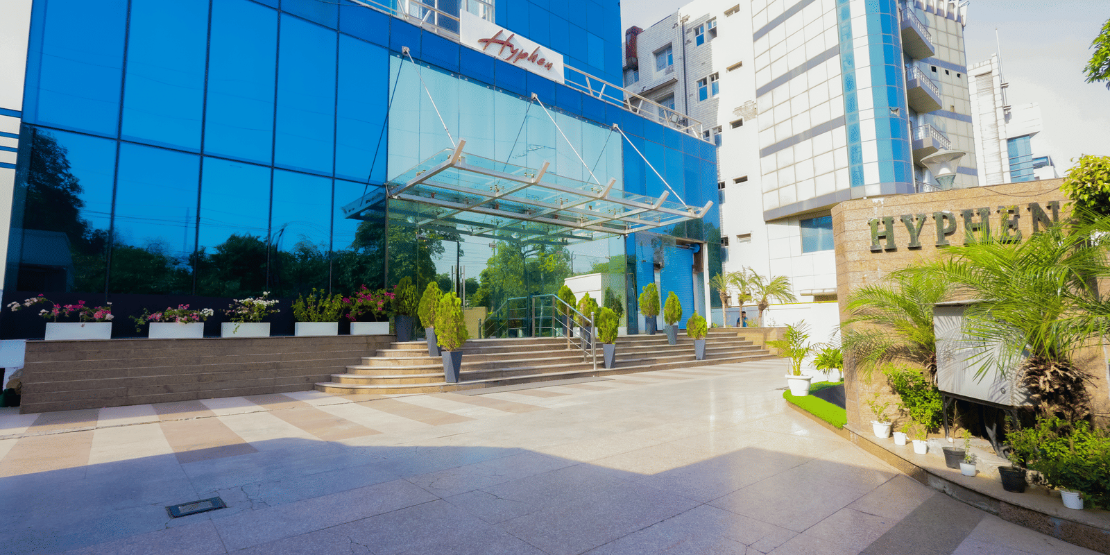 A large, white residential or commercial building with multiple floors stands amidst green landscaping under an overcast sky.