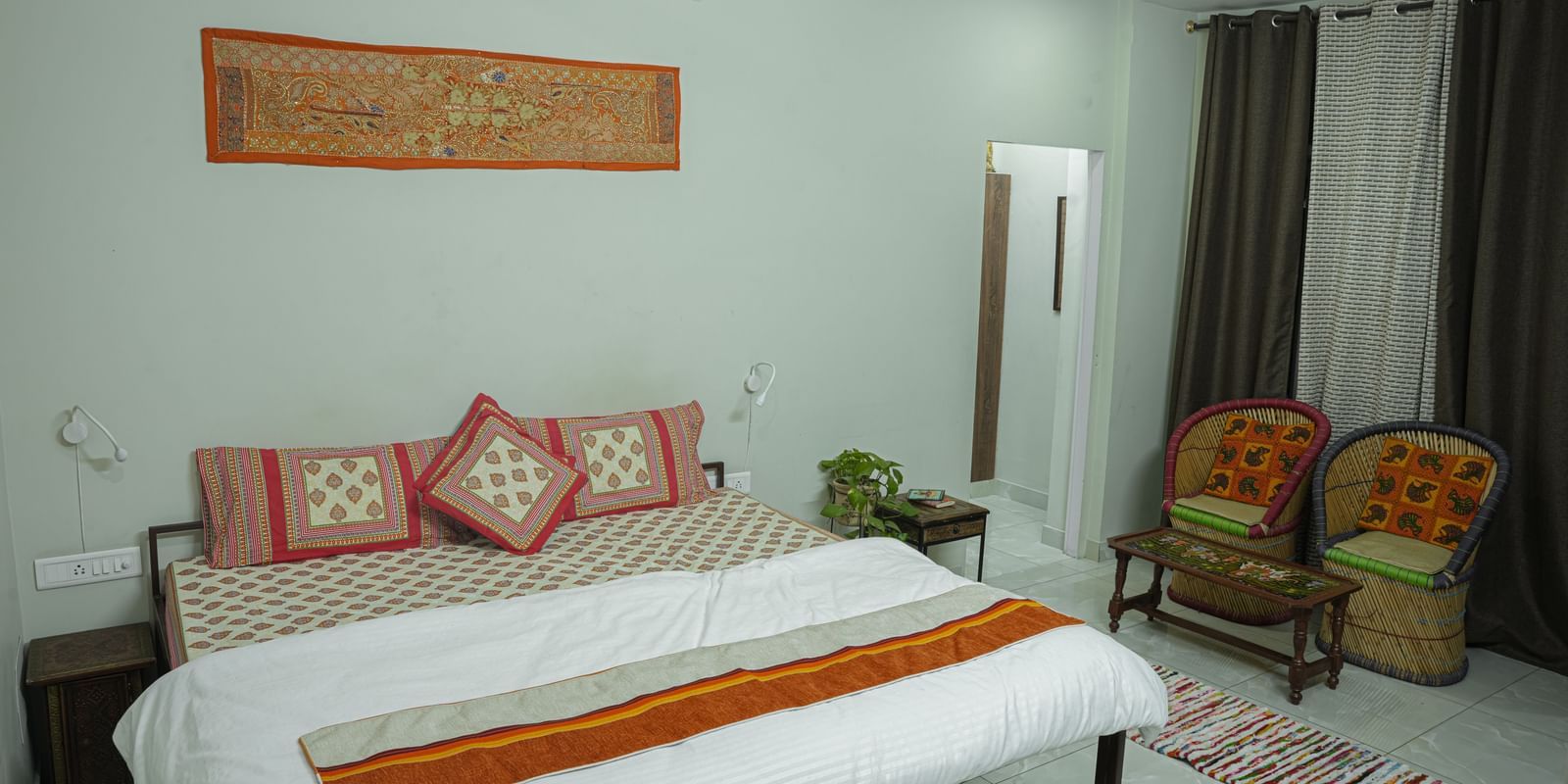 A well-kept bedroom with a double bed, white bedding, orange throw, chairs and wall decor at Jaypore360 Homestay - Best Rooms in Jaipur.