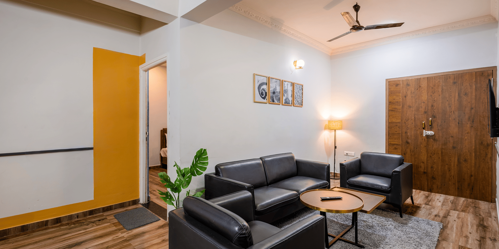 Diagonal view of the separate living room in Coral 4BHK of Kove Smart Stays, Hyderabad, seen with wooden flooring, upholstered multi-seating sofas, a ceiling fan, and a centre table.