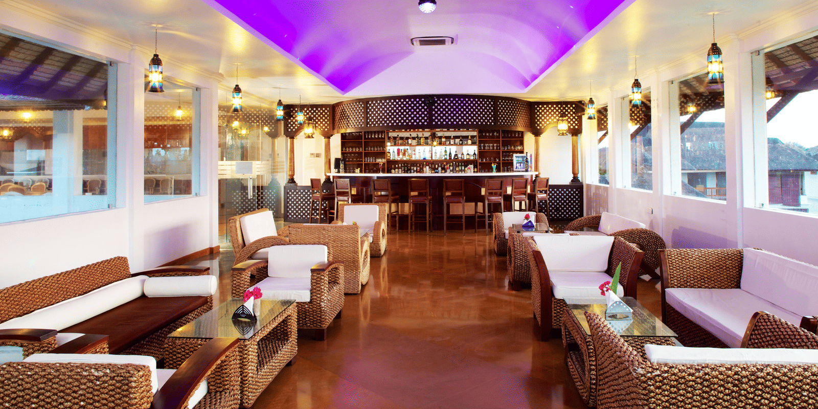 An indoor lounge bar area with wicker sofas, white cushions, and a curved bar counter under a purple-lit ceiling | Le Pondy