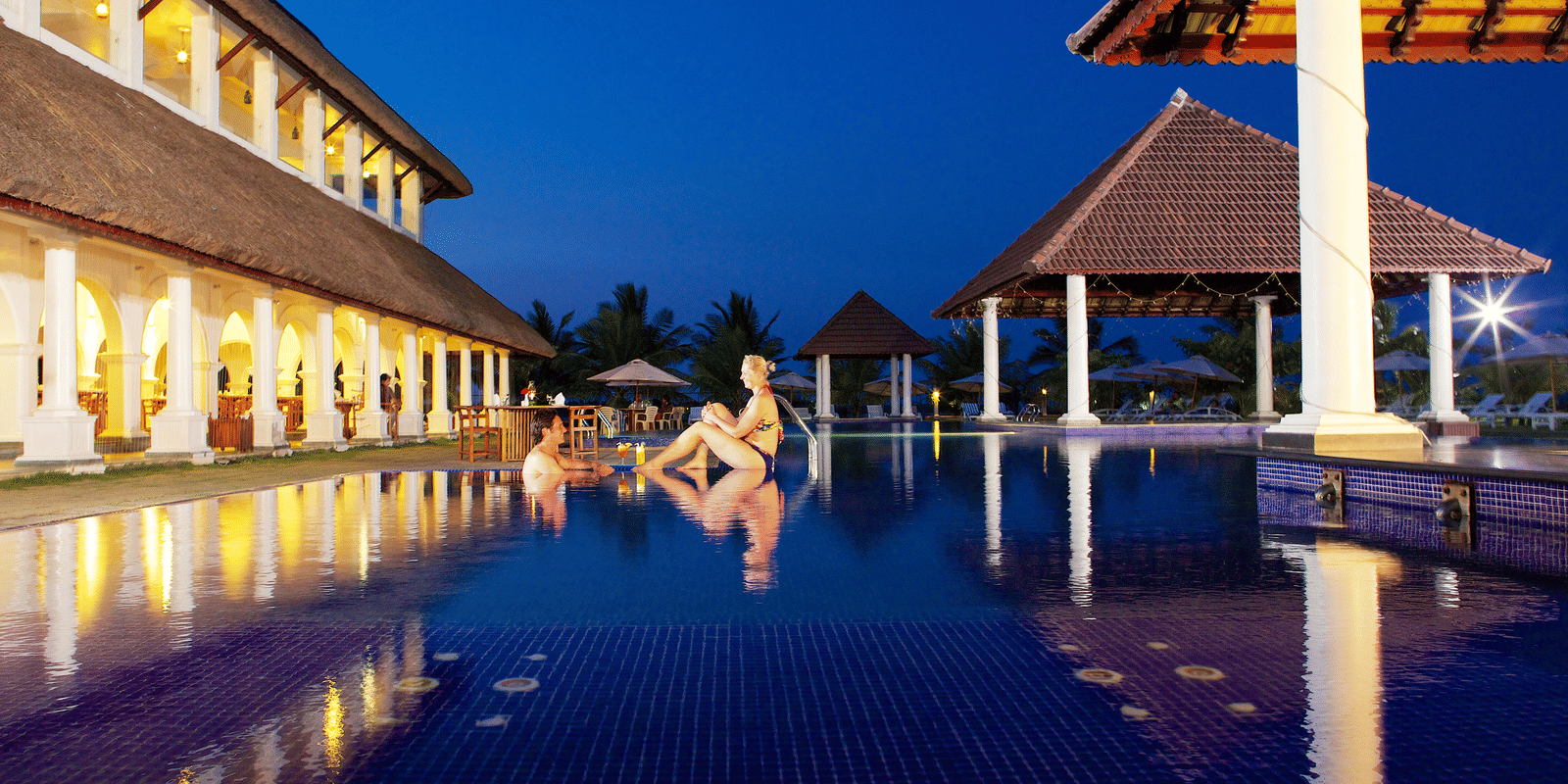 An outdoor swimming pool with blue tiles illuminated at night next to lit resort buildings and pavilions | Le Pondy