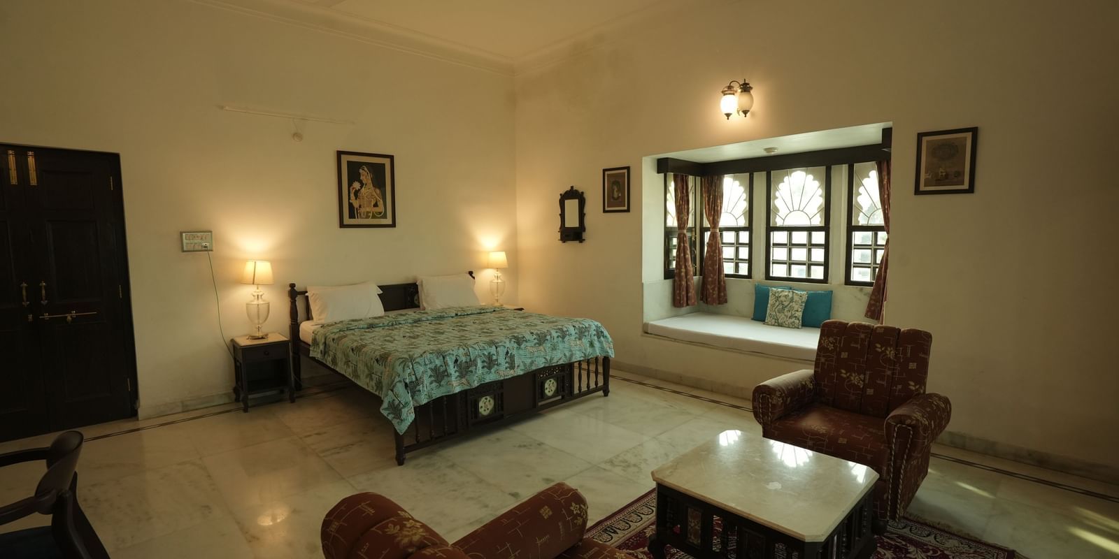 A wide-angle view of the Heritage Courtyard Room at Mahendra Niwas, featuring a large bed, a plush seating area, and traditional windows with curtains.