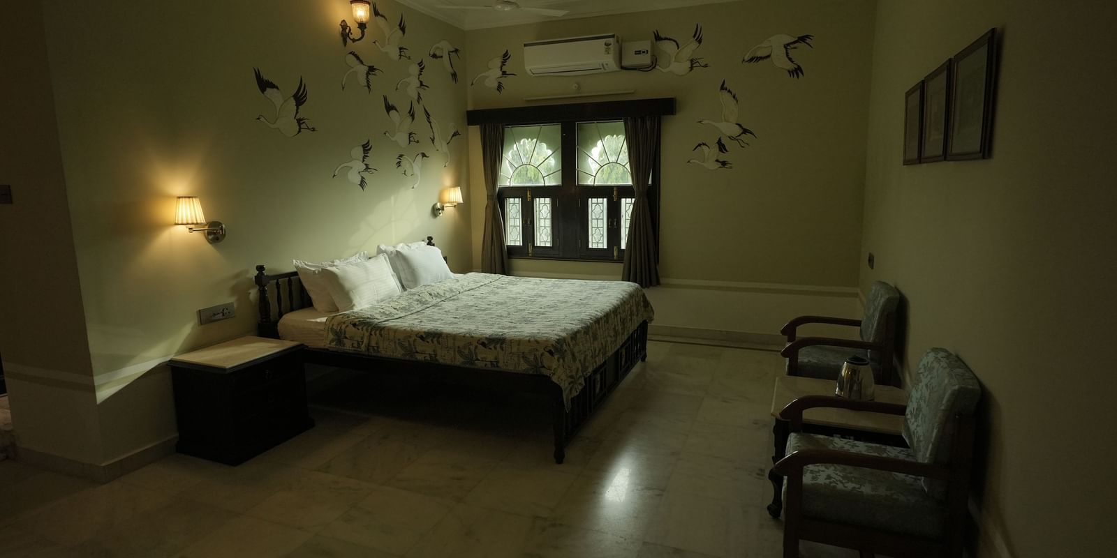 A wide, low-angle shot of the Deluxe Room at Mahendra Niwas. A bed with a light-colored headboard is pushed against a wall decorated with a pattern of black, stylized birds.