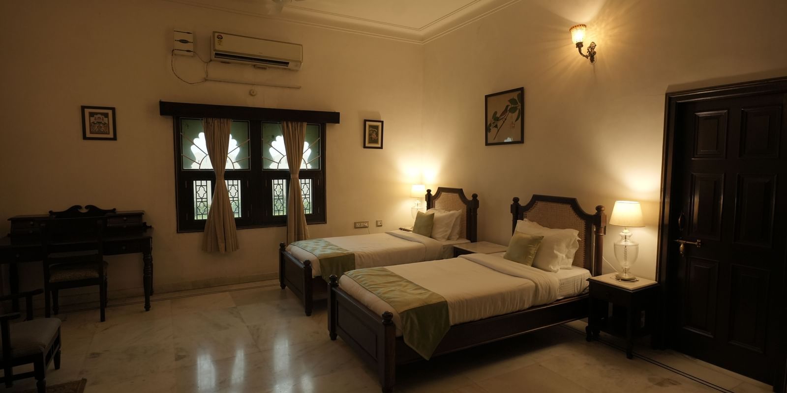 A wide shot of the Terrace Orchard Suite at Mahendra Niwas, showcasing a large room with two twin beds. A small writing desk is visible to the left, and a window looks out onto the grounds.