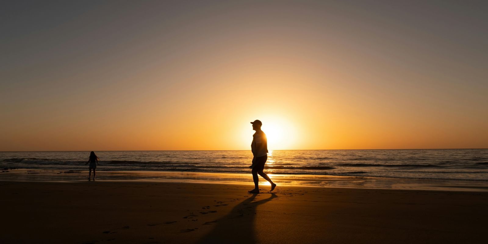 Silhouette of a man walking on a beach side and there is sun setting at the horizon.