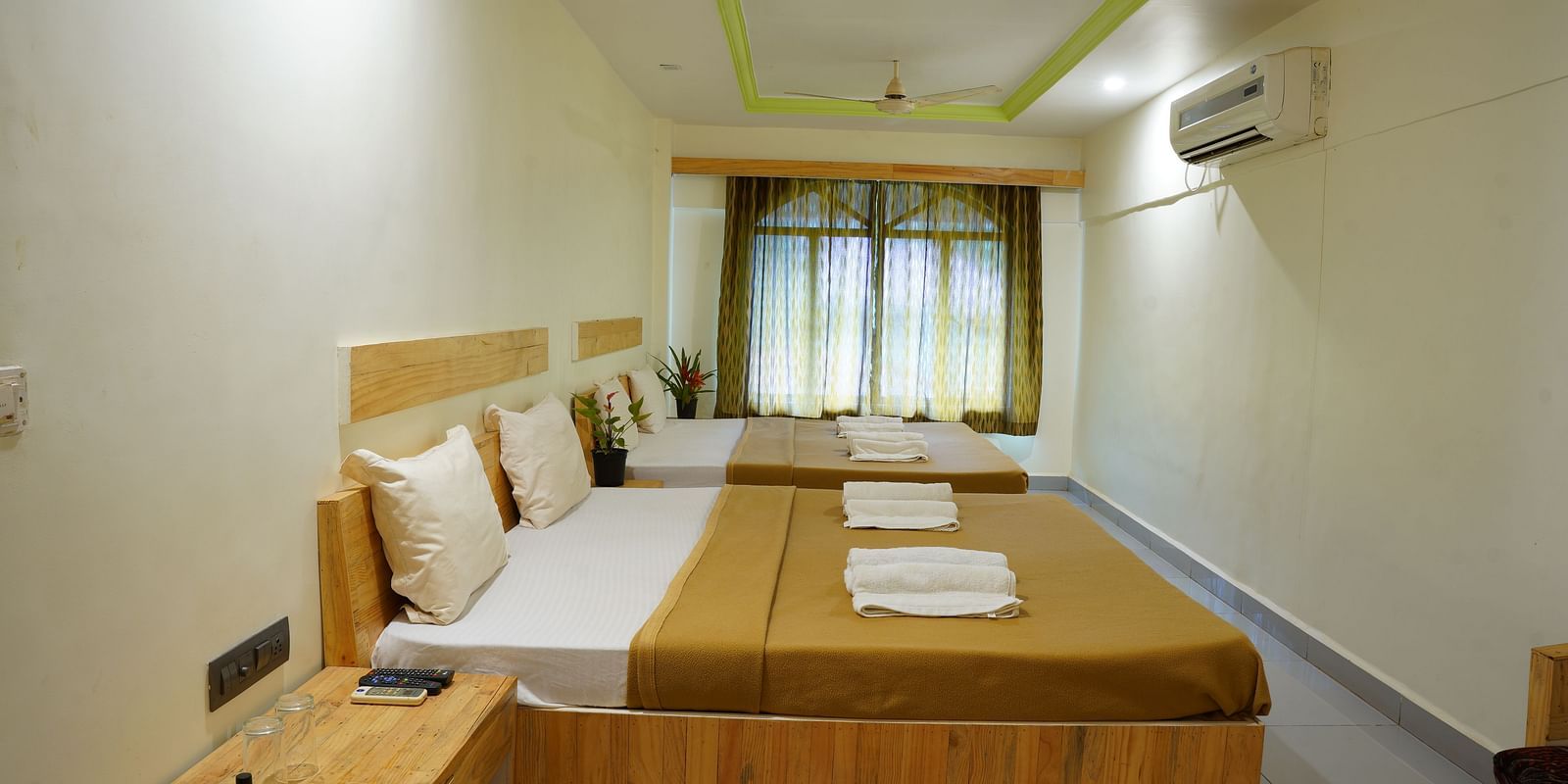 A side view of a room at Riverside Inn, featuring two beds placed beside each other, a bedside table, windows with curtains at the end, and a wall-mounted AC.