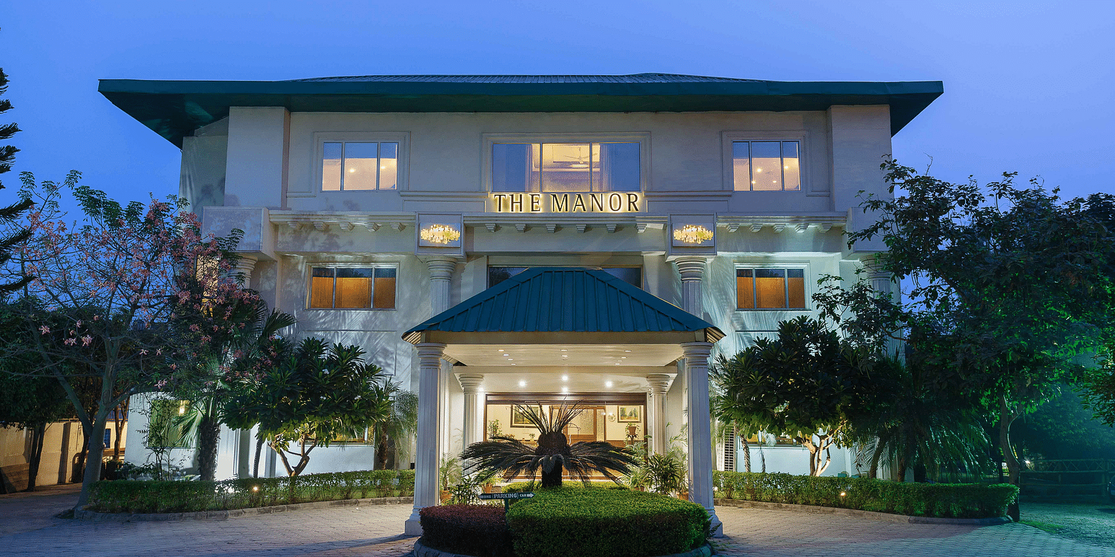A hotel facade framed by lush greenery on both sides, featuring neatly trimmed bushes and a small covered area extends out front, providing a drop-off zone for cars