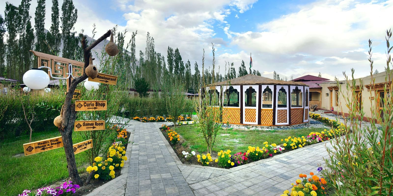 A pathway leading to White Sands Resort, Ladakh lined with flowers and greenery, with decorative signage and accommodation buildings visible against a backdrop of trees and a cloudy sky.
