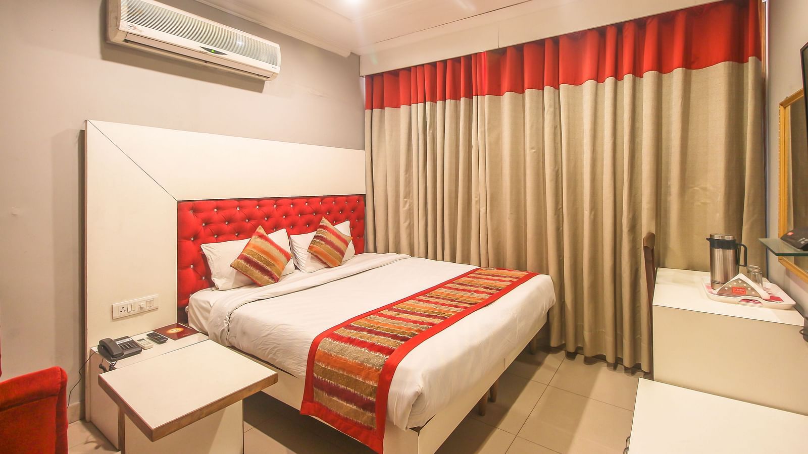 A hotel room with a plush bed featuring a patterned runner, layered curtains, a white headboard, and an AC unit, illuminated by soft lighting. - Hotel Diamond Plaza, Chandigarh.