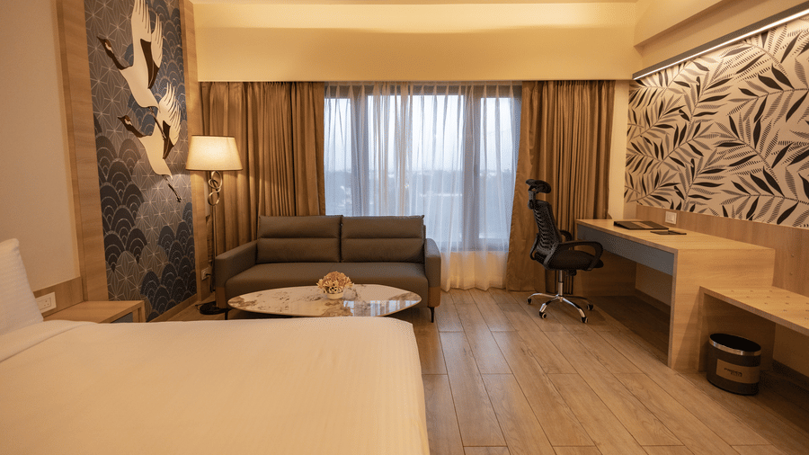 A well-lit hotel room with a large bed in the foreground, a sofa, a work desk, and a decorative wall panel with a leaf pattern | Pride Elite Chhatrapati Sambhajinagar