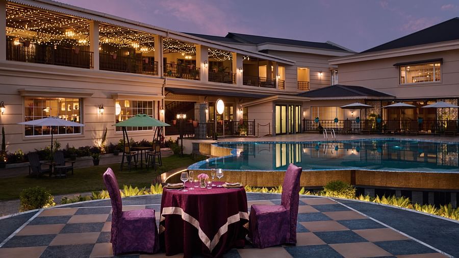 A burgundy-draped round table with two purple chairs sits on a checkered patio, set with wine glasses, plates, and flowers. A lit pool, lounge chairs, and umbrella-covered seating lie beyond, framed by warmly lit multi-story buildings and balcony string li