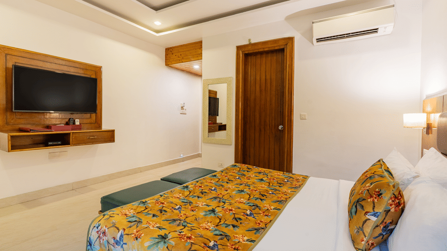 A bedroom featuring a bed with a printed runner, a cushion, a television unit, an A/C unit, and an attached bathroom | Summit by the Ganges Beach Resort & Spa, Rishikesh
