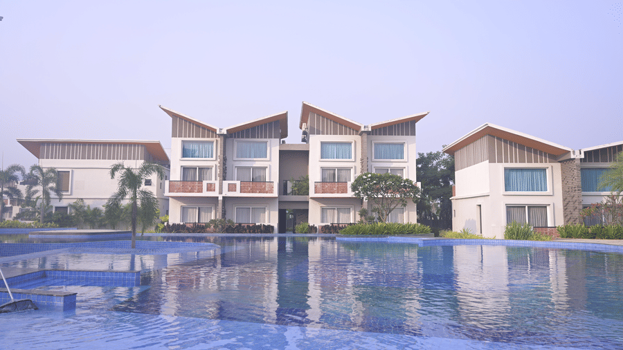 A wide swimming pool reflecting the hotel buildings under a clear sky at Hotel Sonar Bangla, Mayapur.