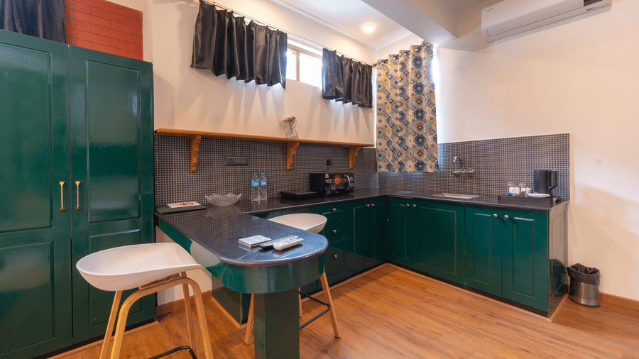 A contemporary studio kitchenette with dark green cabinetry, wooden flooring, and a breakfast bar with a white stool - Johnson Lodge & Spa, Manali