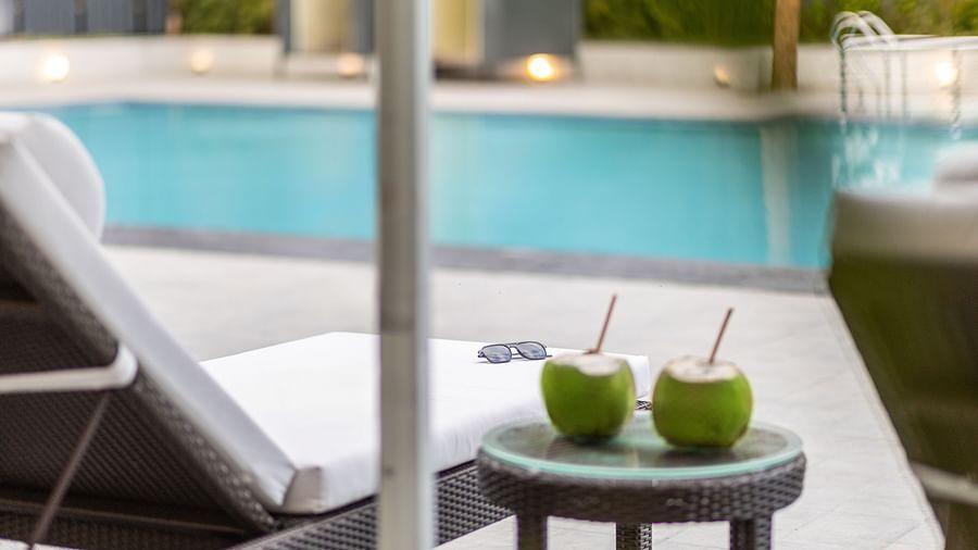 A poolside scene with a lounge chair, two coconuts on a table, and a swimming pool in the background - Mango Hill Resort, Coimbatore