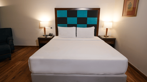 A room at Park Plaza Ludhiana featuring a king bed with white bedding and teal checkered headboard wooden flooring bedside tables with lamps and warm ambient lighting.