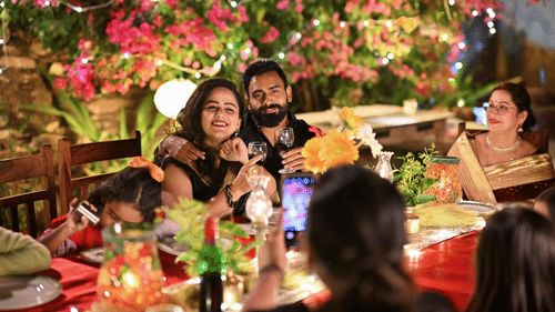 A couple holding a glass of wine in a cocktail soiree where other people have also joined. The setting is amidst a garden lit with flowers and fairy lights. 