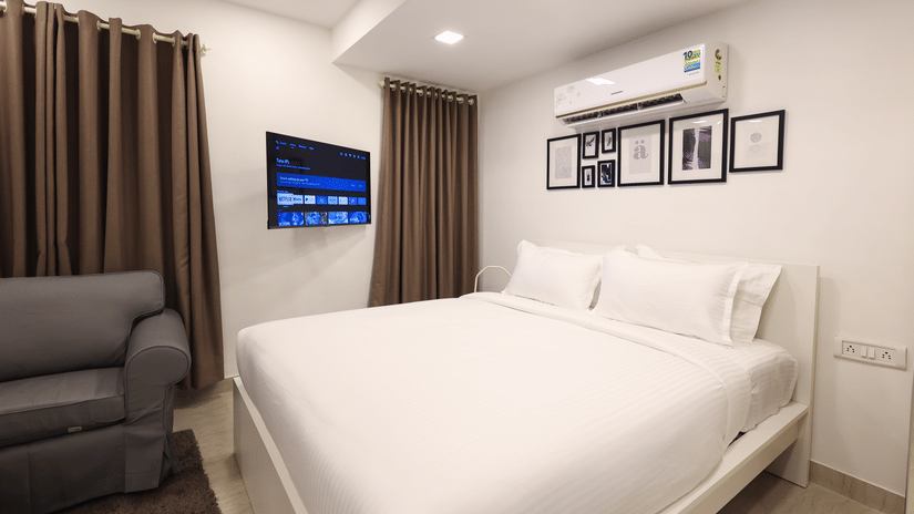 A bedroom at Wabi - Sabi by Alamiko, Juhu, featuring a plush bed, a sofa, framed painting on a wall, an A/C unit, and a smart TV.
